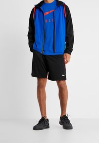 Man i svarta sportshorts med neonlogga, blå grafisk T-shirt, blå och svart zip-up jacka samt svarta löparskor som står.
