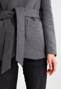 Abrigo de mezcla de lana gris con un cinturón ancho, dos bolsillos frontales y bordes de bolsillo curvados. Lleva puesto sobre pantalones negros, mostrando la textura de la tela.