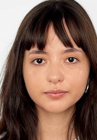 Zbliżenie osoby z długimi, ciemnobrązowymi włosami i jasną cerą. Cechy to proste grzywki, gładka cera i subtelny makijaż.