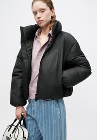 Čierna nadýchaná bunda s vysokým golierom, vybavená zipsom. Model má na sebe textúrovanú ružovú košeľu a svetlo modré pruhované džínsy, v ruke drží bielu tašku.