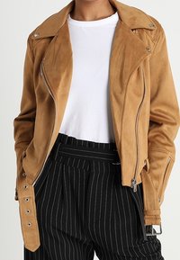 Veste de moto en daim tan avec des fermetures éclair argentées et des boutons-pression, présentant un devant asymétrique et des coutures détaillées. Associée à un pantalon rayé.