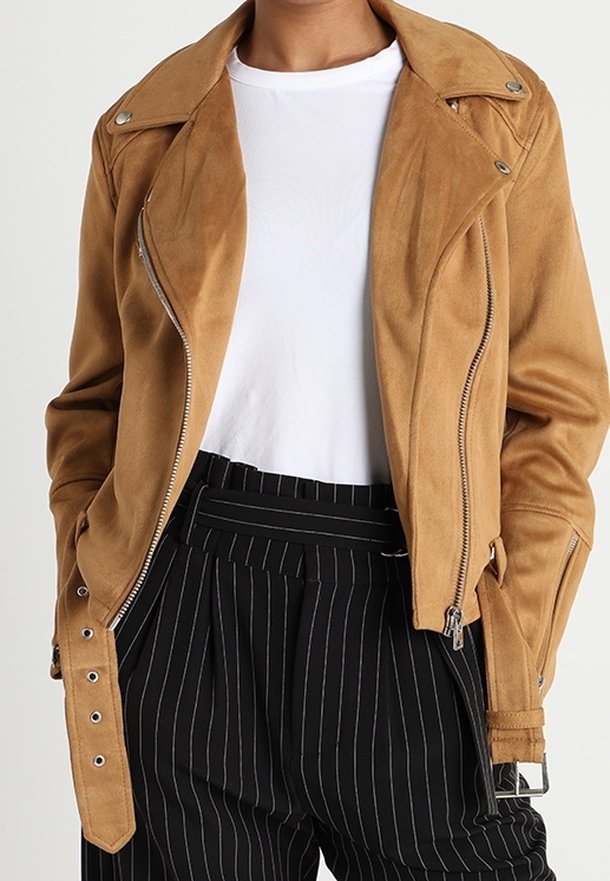 Veste de moto en daim tan avec des fermetures éclair argentées et des boutons-pression, présentant un devant asymétrique et des coutures détaillées. Associée à un pantalon rayé.