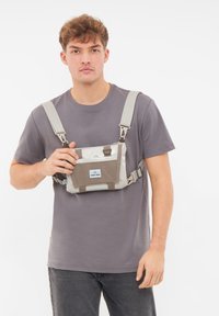 Borsa a tracolla grigia e beige con cinturini regolabili, tasca frontale con zip e dettaglio del logo. Il materiale sembra durevole e impermeabile.