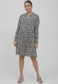 Schwarz-weiß gemustertes Kleid mit langen Ärmeln, knöpfbarem Kragen und gestuftem Rockdesign; kombiniert mit hellgrauen Absatzsandalen.