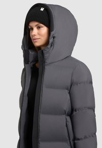 Chaqueta acolchada gris con una capucha grande, que presenta un cierre de cremallera, un exterior liso y una textura acolchada. Incluye un gorro de punto negro.