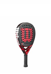Wilson padel racket met een zwarte oppervlakte, groot rood logo, perforaties voor grip en een gestructureerde handgreep afgewerkt in zwart rubber.