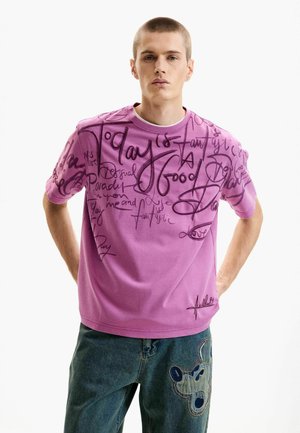 Camiseta rosa de manga corta con texto y diseños en negro estilo manuscrito. Tela suave con escote redondo, combinada con pantalones vaqueros azules.