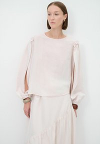 Blusa rosa chiaro con maniche lunghe a sbuffo e fiocchi sulle spalle. Realizzata in tessuto morbido e traslucido, con una vestibilità morbida e scollatura rotonda.