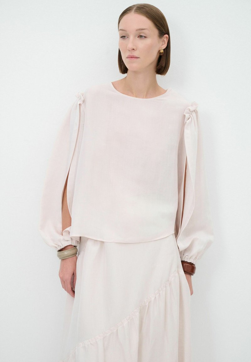Blusa rosa chiaro con maniche lunghe a sbuffo e fiocchi sulle spalle. Realizzata in tessuto morbido e traslucido, con una vestibilità morbida e scollatura rotonda.
