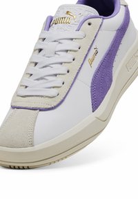 Tenis cu piele albă, cu accente din suede mov, talpă medie crem și logo Puma aurie. Prezintă un design cu șireturi și detalii texturate pe limbă.
