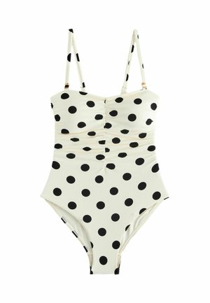 Costum de baie întreg, alb, cu buline negre și bretele subțiri reglabile pe un fundal simplu.