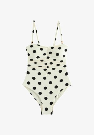 Bijeli jednodijelni kupaći kostim s crnim točkicama i tankim podesivim remenima na ramenu na jednobojnoj pozadini.