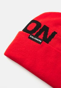 Rode gebreide muts met een omslag, voorzien van grote zwarte "SALOMON" tekst en een klein zwart label. Gladde textuur, warm materiaal.