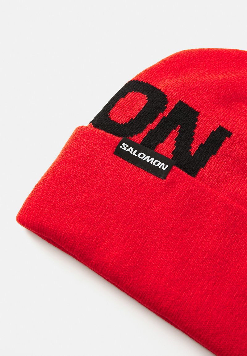 Rode gebreide muts met een omslag, voorzien van grote zwarte "SALOMON" tekst en een klein zwart label. Gladde textuur, warm materiaal.