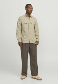 Camicia button-up beige chiaro con due tasche sul petto, indossata sopra un top color tan. Abbinata a pantaloni marroni larghi e sneakers beige.
