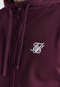 Gros plan sur un sweat à capuche zippé bordeaux avec cordons assortis et un logo brodé blanc orné sur le côté gauche de la poitrine.