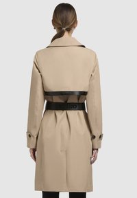 Beige trench coat with a black belted waist, large collar, and buttoned cuffs. Back features a black logo detail and a streamlined silhouette.