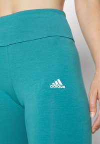 Kvinnors teal atletiska leggings tillverkade av ett smidigt, stretchigt tyg med en hög midja och en vit Adidas-logotyp på vänster lår.