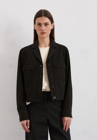 Zwarte cropped jas met een kraag, knopen aan de voorkant, twee borstzakken en lange mouwen. Gedragen over een lichtgekleurde trui en donkere broek.