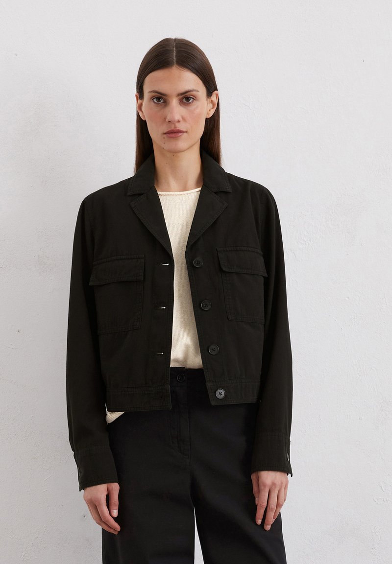Zwarte cropped jas met een kraag, knopen aan de voorkant, twee borstzakken en lange mouwen. Gedragen over een lichtgekleurde trui en donkere broek.