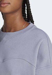 Primer plano de una persona con cabello trenzado oscuro que lleva un sudadera de cuello redondo en un tono lila claro y un collar delgado de oro.