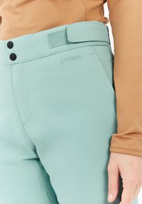 Lichtblauwe broek met een gladde textuur, voorzien van twee zwarte knopen, een klittenband tailleband en een subtiele "protest" logo aan de voorkant.