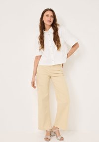 Weißes, kurzes Button-up-Hemd mit kurzen Ärmeln, kombiniert mit beigen, weiten Hosen und silbernen, hochhackigen Sandalen. Glatte Texturen überall.