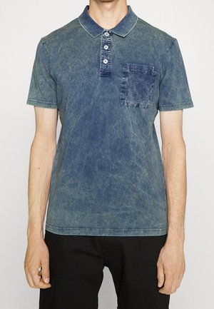 Mann trägt ein ausgebleichtes blaues Polohemd mit Knopfleiste und Brusttasche, kombiniert mit schwarzen Hosen, vor einfachem Hintergrund.