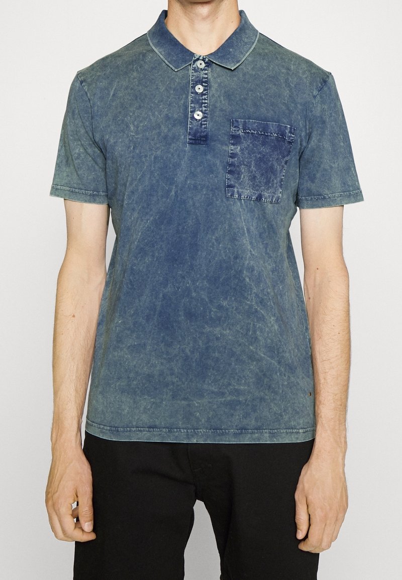 Homme portant un polo bleu délavé avec patte de boutonnage et poche poitrine, associé à un pantalon noir, sur un fond uni.