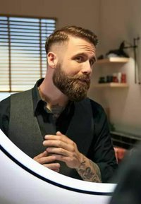 Mann mit einem kurzen Bart und Tattoos, der ein schwarzes Shirt und eine graue Weste trägt, steht an einem runden Spiegel mit beleuchtetem Rand und pflegt sich.