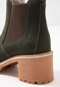 Botín de piel verde con panel elástico marrón, tacón texturizado color miel y detalle de costura contrastante a lo largo de las costuras.