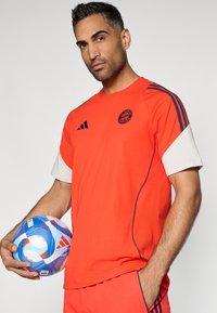 Piros sportpóló fehér ujjal, kékkal és fehérrel mintázott focilabdát tart a modell.