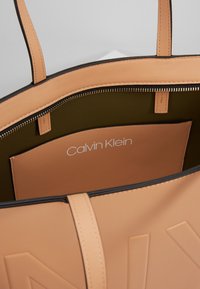 Kabelka Calvin Klein z měkké béžové kůže, s olivově zeleným interiérem, zipovým uzávěrem a embosovaným logem na přední kapse.