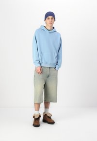 Hanorac cu glugă de culoare bleu deschis, pantaloni scurți din denim verde deschis, cizme maro cu terminatie din blană sintetică și o șapcă tricotată bleu. Îmbrăcăminte casual.