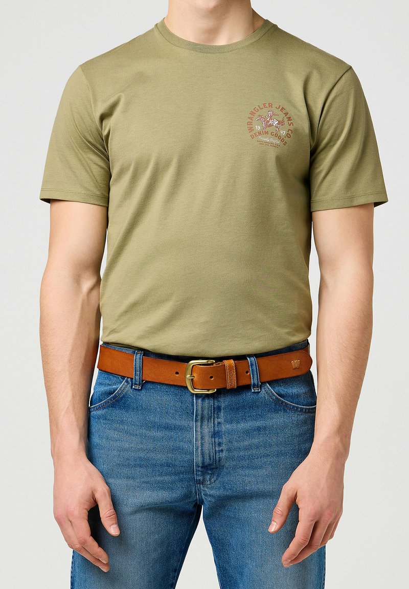 Olivgrünes Baumwoll-T-Shirt mit kurzen Ärmeln und Rundhalsausschnitt, mit einem bedruckten Logo auf der Brust. Getragen mit einem braunen Ledergürtel und blauen Jeans.