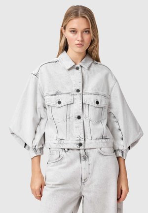 Młoda kobieta w jasnoszarej kurtce jeansowej typu acid wash z bufiastymi rękawami oraz dopasowanych, wysokich spodniach jeansowych, na jednolitym tle.