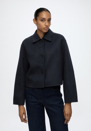 Vrouw met donker haar in een staart, een zwarte jas met knopen en donkere spijkerbroek, staand voor een effen witte achtergrond.