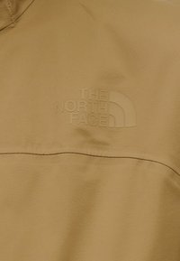 Μπεζ εξωτερικό μπουφάν με λεία υφή και εμφανές ανάγλυφο λογότυπο "THE NORTH FACE" στην αριστερή πλευρά.