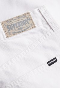 Tissu blanc à surface texturée. Comprend une grande étiquette en papier embossée du mot « SUPERDRY » et une petite étiquette noire avec « SUPERDRY ».