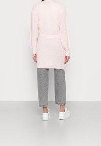 Světle růžový dlouhý pletený cardigan s texturovaným vzorem, volným střihem a žebrovanými manžetami, doplněný o šedé volné kalhoty a bílé tenisky.