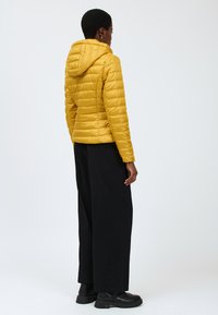 Gul pufferjakke med hætte, quiltet design og slim fit. Båret med sorte vide bukser og ankelstøvler, hvilket viser en glat tekstur.