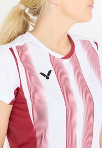 Punainen ja valkoinen urheilupaita, jossa on kapeat pystysuorat raidat, pyöreä pääntie ja ympyrälogo etupuolella. Valmistettu hengittävästä materiaalista.