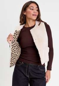 Femme portant un gilet en fausse fourrure crème par-dessus une chemise à manches longues marron, montrant la doublure imprimée léopard à l'intérieur du gilet.