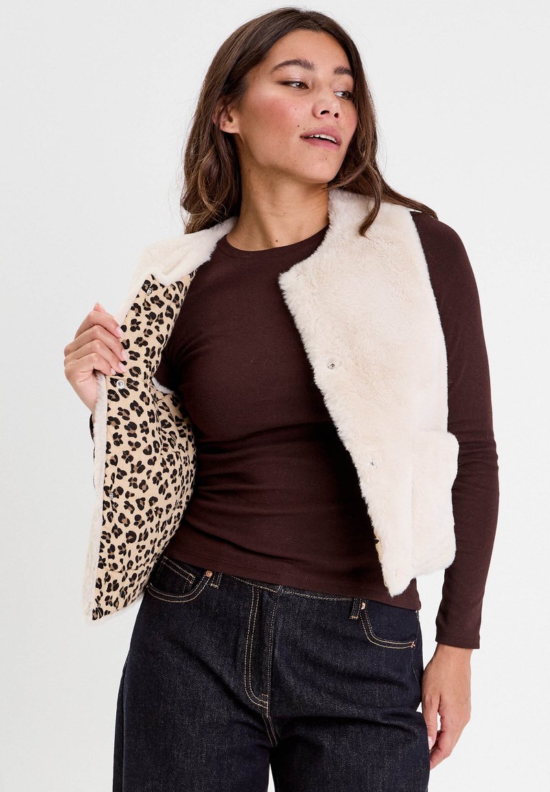 Femme portant un gilet en fausse fourrure crème par-dessus une chemise à manches longues marron, montrant la doublure imprimée léopard à l'intérieur du gilet.