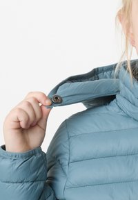 Ljusblå puffjacka med hög krage, med en synlig tryckknapp och vadderad textur. Ärmen har en mudd med en slät yta.