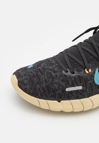 Baskets en tricot noires avec semelle beige, logo bleu sur le bout et sur le côté, lacets noirs et boucles de lacets jaunes.