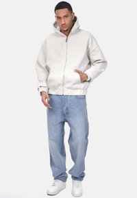 Haut de hoodie zippé gris clair avec une capuche doublée, des poignets élastiques et des poches latérales, associé à un jean bleu clair ample et des baskets blanches.