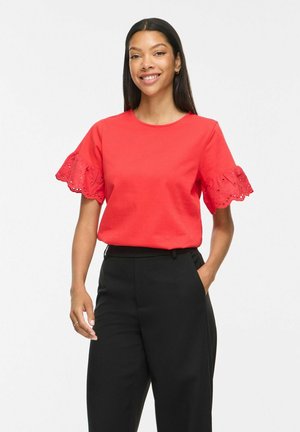 Femme souriante aux cheveux longs et raides, portant un haut rouge avec des manches à volants en dentelle et un pantalon noir taille haute, debout avec une main dans la poche.