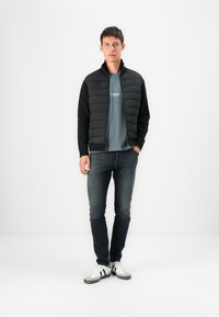 Black puffer jacket with a zip closure and ribbed cuffs, paired with a gray t-shirt, dark jeans, and white sneakers with black stripes.