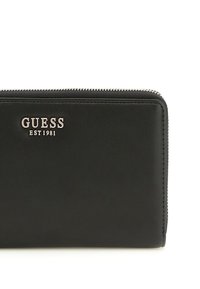 Portafoglio in pelle nera con chiusura a zip, caratterizzato da un logo metallico "GUESS EST 1981" sulla parte anteriore, superficie liscia e forma rettangolare.